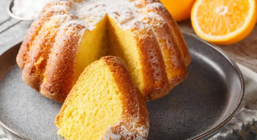 Puszysta babka na oleju, oprószona cukrem pudrem, z pomarańczami w tle. Idealna na niedzielne popołudnie.