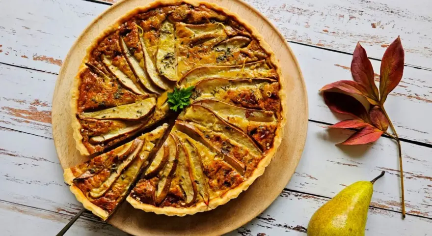 Apetyczna tarta z gruszką i serem pleśniowym, ozdobiona natką pietruszki i skórką cytrynową, na drewnianej desce. Obok leży gruszka i jesienne liście.