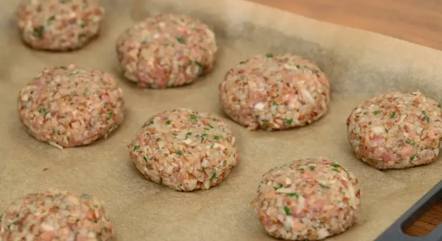 Surowe burgery z piekarnika, przygotowane z mielonego mięsa i kaszy, leżą na papierze do pieczenia.