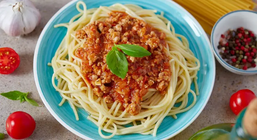 Pyszny, prosty sos do spaghetti z mielonym mięsem i pomidorami, udekorowany bazylią. Obok czosnek, pomidorki i kolorowy pieprz.