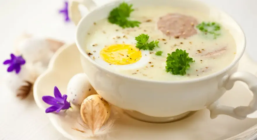 Aromaticzny barszcz biały na żeberkach z jajkiem i kiełbasą, ozdobiony natką pietruszki. Na talerzyku wielkanocne jajka i fioletowe kwiaty.