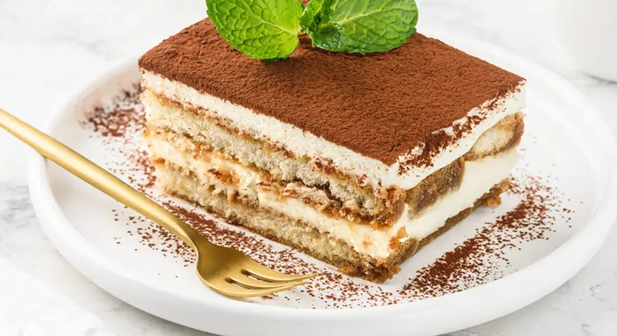 Pyszne tiramisu z mascarpone i śmietaną, posypane kakao i ozdobione miętą. Złoty widelec czeka na pierwszy kęs.