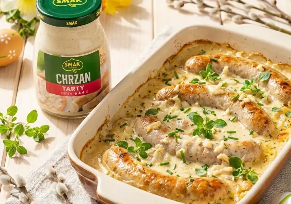 Biała kiełbasa pieczona w kremowym sosie chrzanowym, ozdobiona ziołami. Obok słoik sosu chrzanowego.