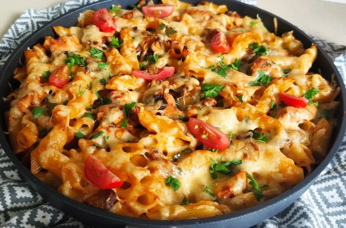 Pyszna zapiekanka z kurczakiem, makaronem penne, roztopionym serem i pomidorkami koktajlowymi, posypana natką pietruszki.