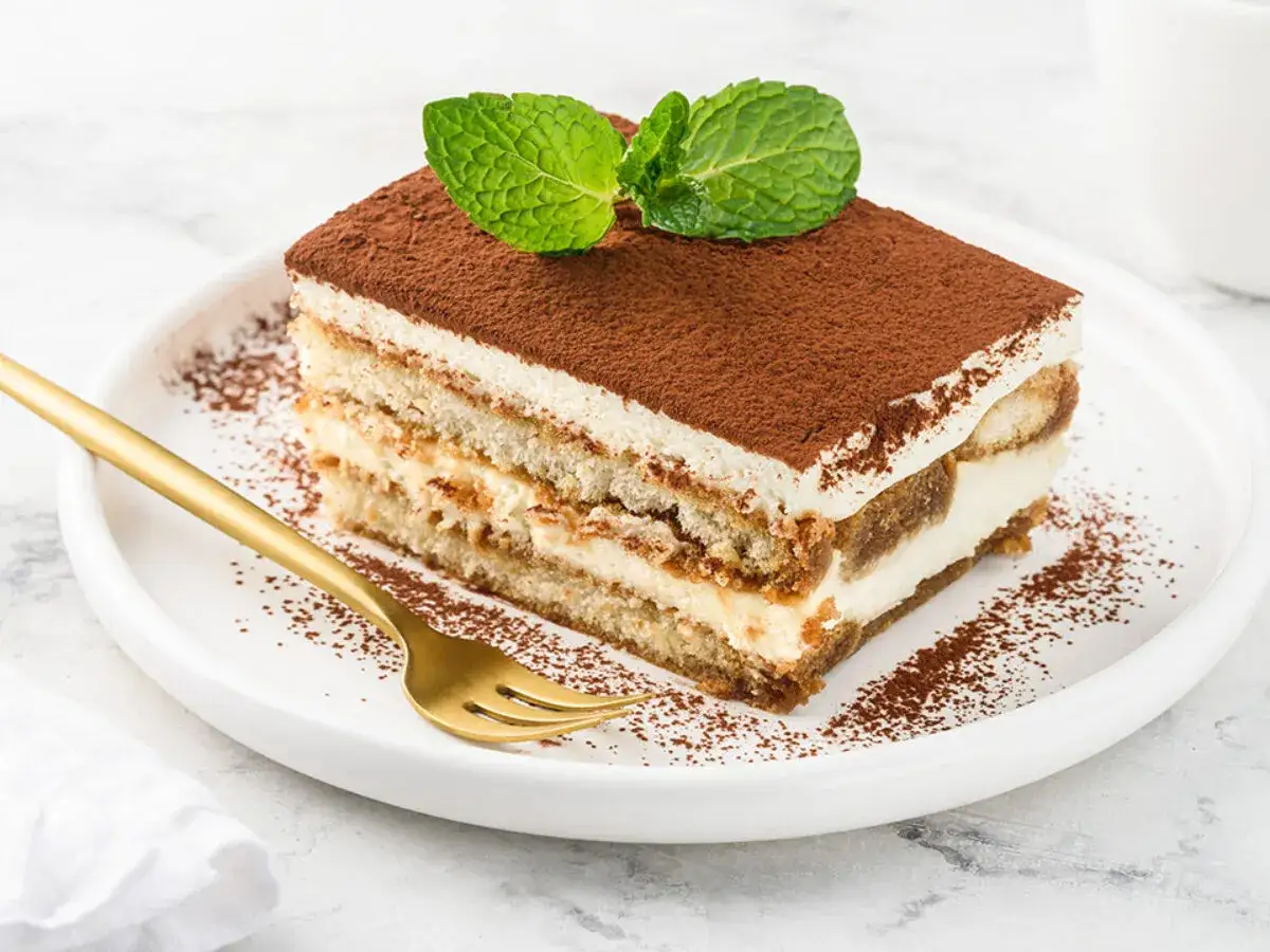 Pyszne tiramisu z mascarpone i śmietaną, posypane kakao, ozdobione listkami mięty. Złoty widelec czeka na pierwszy kęs.