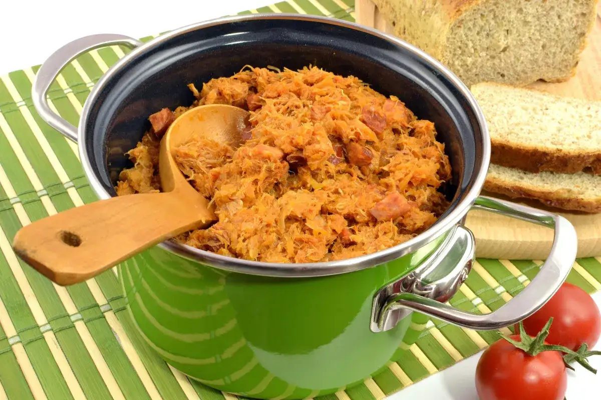 Gotowy bigos w zielonym garnku, obok kromki chleba i pomidory. Idealne przyprawy do bigosu sprawiają, że danie wygląda apetycznie.