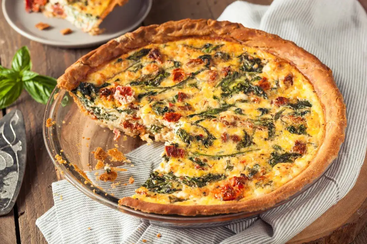 Pyszna tarta ze szpinakiem i suszonymi pomidorami, z kawałkiem już wyjętym. Idealna na lunch lub kolację.