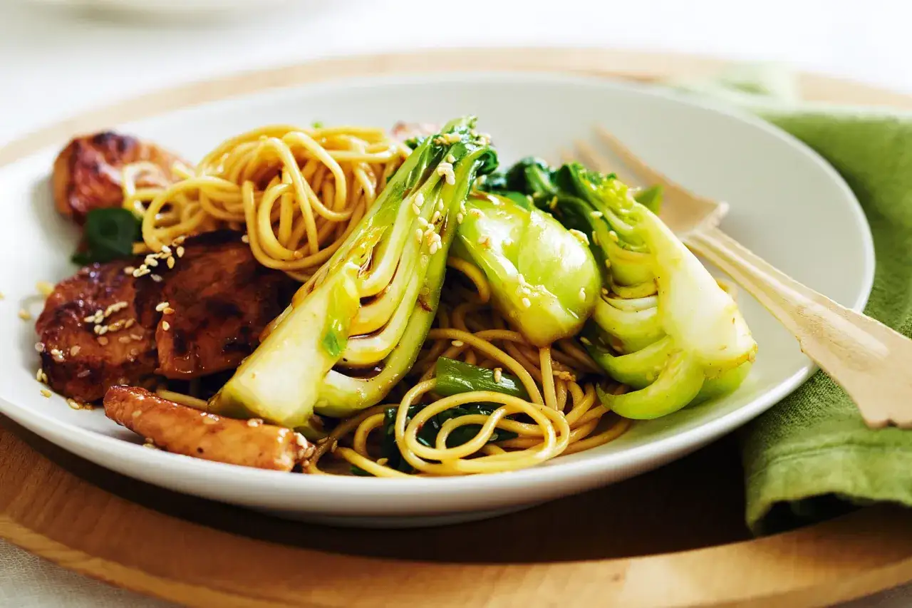 Apetyczny kurczak teriyaki z makaronem i pak choi, posypany sezamem. Danie podane na białym talerzu, obok zielona serwetka i drewniane sztućce.