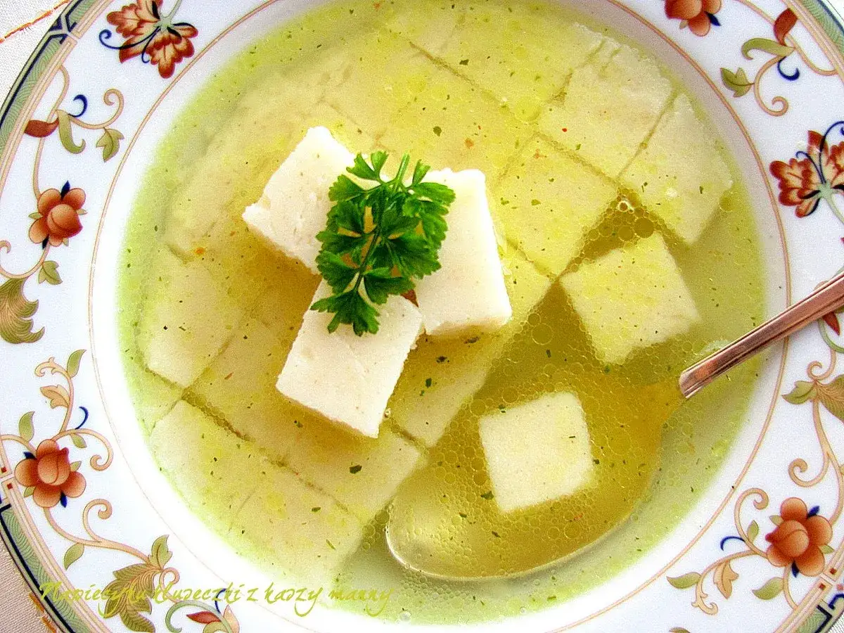 Zupa z kaszą manną, podana w ozdobnej miseczce, z kostkami kaszy i gałązką natki pietruszki.