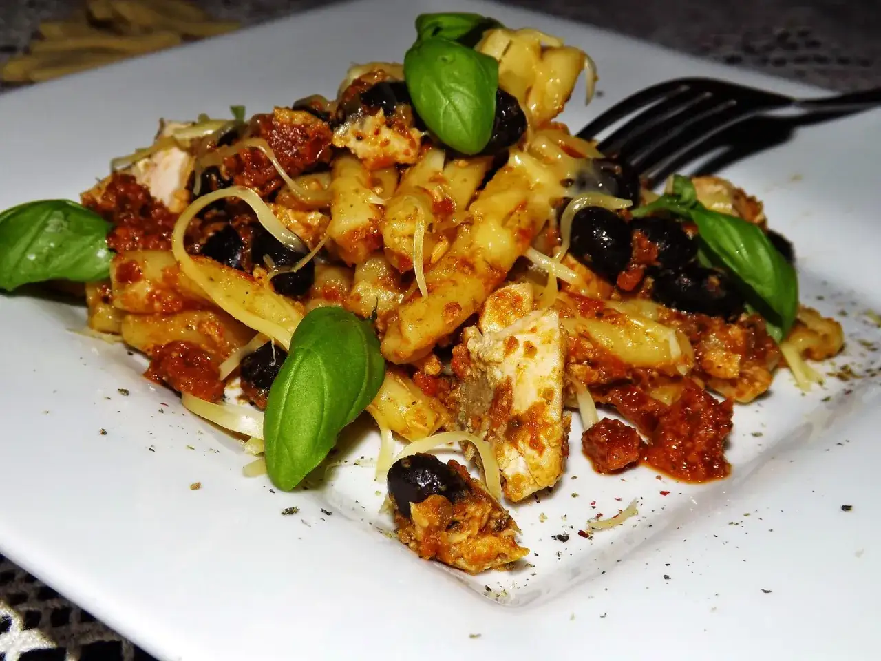 Penne z tuńczykiem, czarnymi oliwkami i suszonymi pomidorami, polane sosem pesto, ozdobione listkami bazylii.