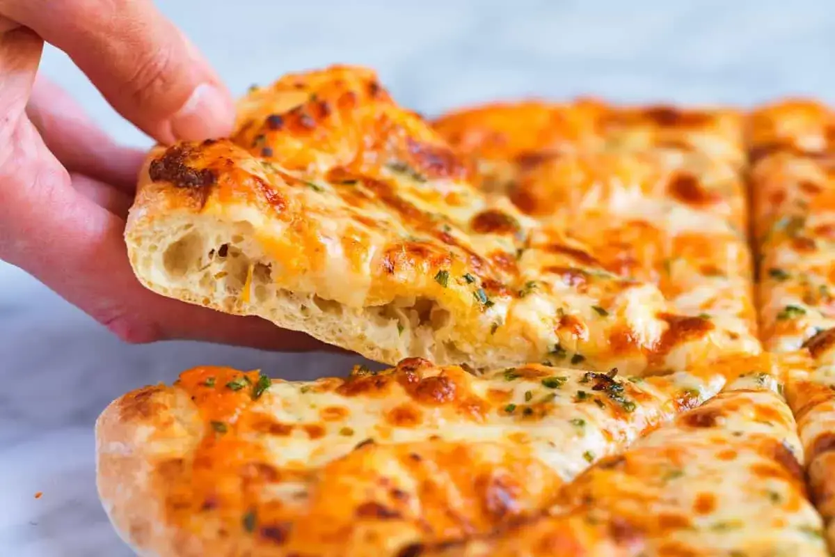 Ręka podnosi kawałek puszystego ciasta na pizzę, obficie posypanego serem i ziołami.