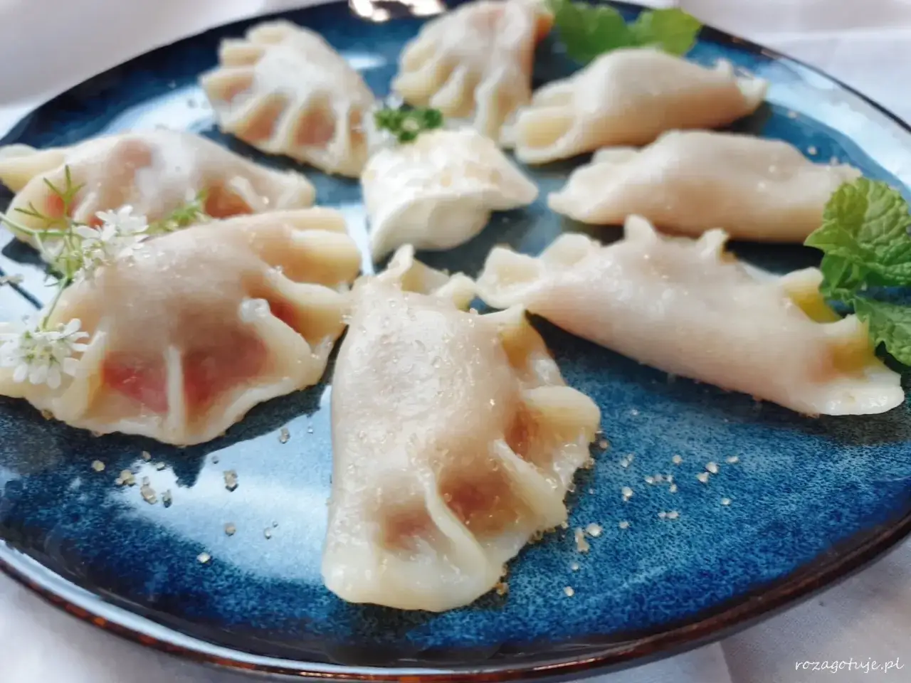 Pyszne pierogi z wiśniami, ozdobione listkami mięty i drobnymi kwiatkami, kuszą na niebieskim talerzu.