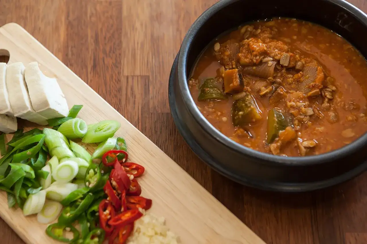 Gulasz z kaszą w misce, obok deska z pokrojonym tofu, szczypiorkiem, papryczkami chili i czosnkiem.