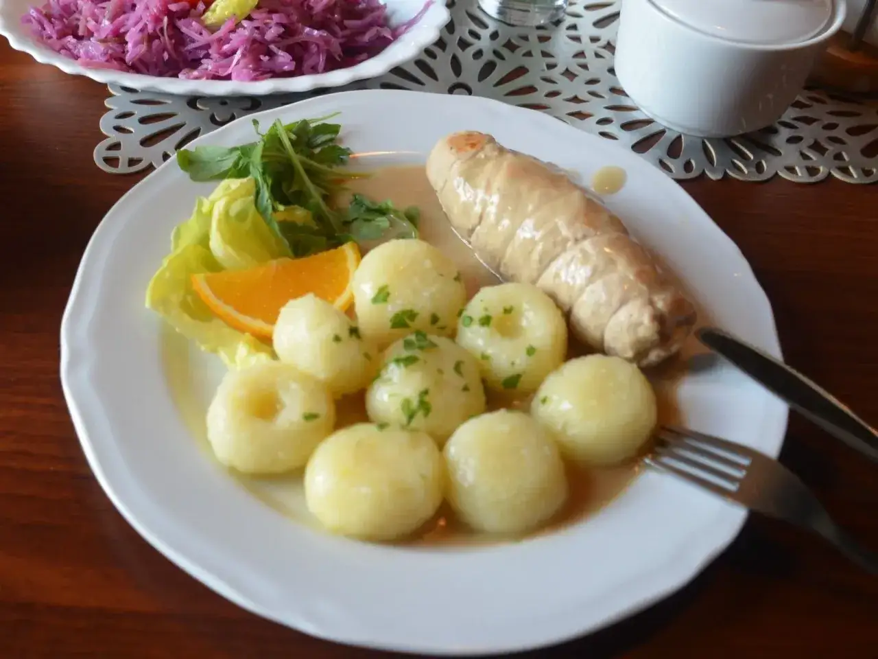 Jak zrobić kluski śląskie? Na talerzu rolada z mięsa, kluski śląskie z koperkiem, plaster pomarańczy i sałatka z czerwonej kapusty.
