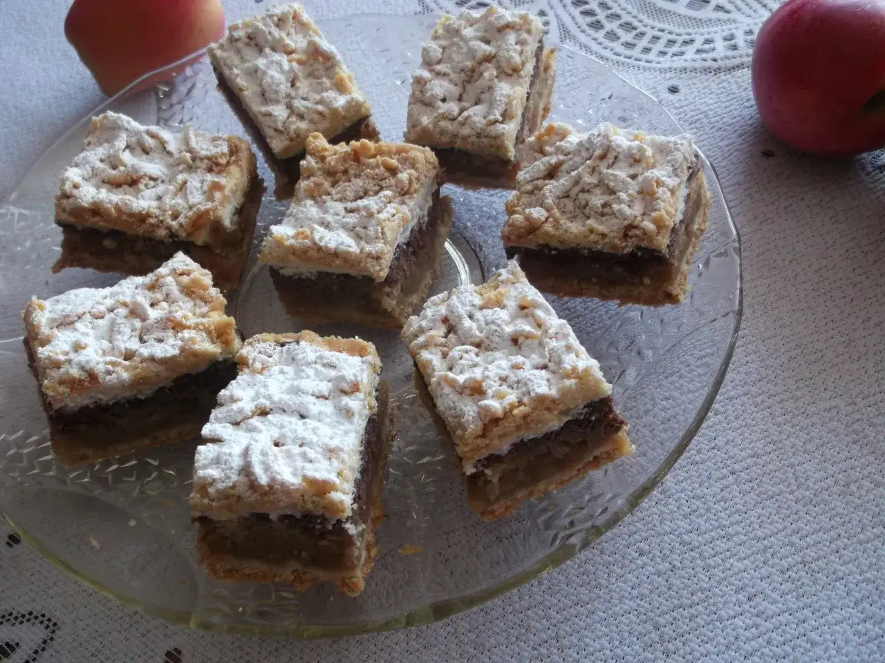 Pyszny pleśniak z jabłkami, posypany cukrem pudrem, na szklanej paterze. Obok leżą jabłka.