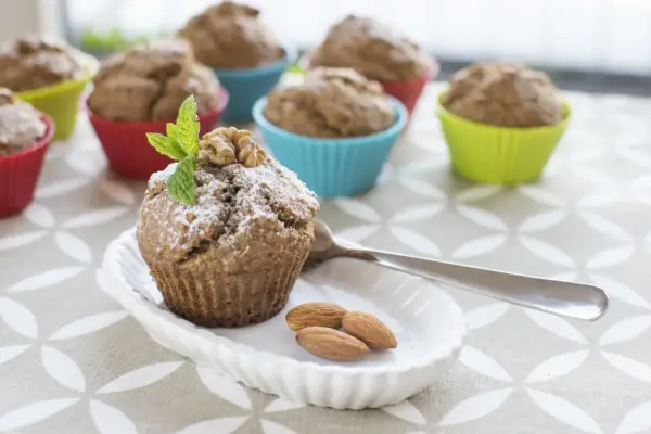 Pyszne muffinki bananowe fit, posypane cukrem pudrem, z orzechami i miętą. Obok migdały i łyżeczka.