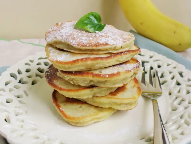 Stosik puszystych placuszków bananowych bez mleka, posypanych cukrem pudrem, z bananem w tle.